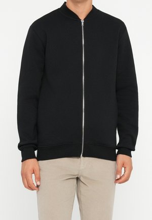 Homme portant une veste noire zippée avec poignets et ourlet côtelés, associée à un pantalon beige clair, debout devant un fond uni.