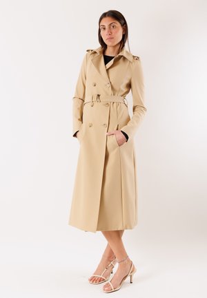 Donna in piedi con le mani nelle tasche, indossa un trench color beige con cintura e sandali beige con tacco e punta aperta, su uno sfondo neutro.