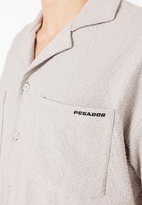 Vaaleanharmaa, teksturoitu paita, jossa on viistokaulus, etutasku ja musta "PEGADOR" -kirjailu. Painike sulkeminen ja pehmeä kangas.