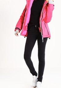 Doudoune isolante rose avec des accents noirs, portée sur un pull noir, associée à un jean skinny noir et des baskets blanches.