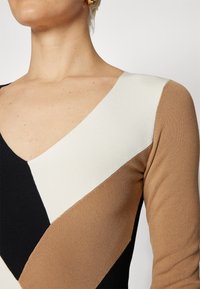 Femme portant un haut à col en V avec des blocs de couleur diagonaux en noir, blanc et marron, montrant les détails du cou, de l'épaule et du haut de la poitrine.