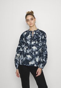 Blusa azul-marinho e branca com padrão, de decote redondo e detalhe em buraco. Apresenta mangas longas e bufantes, e um corte relaxado.