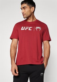 Rød bomull T-skjorte med "UFC"-logo og grafikk, korte ermer, rund hals og avslappet passform. Kombinert med svarte bukser.