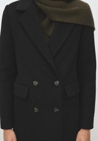 Czarny dwurzędowy płaszcz z dużymi guzikami i dwoma przednimi kieszeniami, noszony na teksturowanym, ciemnozielonym, dzianym szaliku.