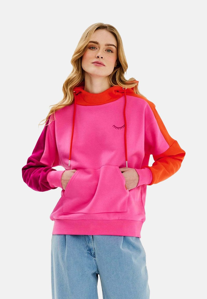 Vibrant růžová a oranžová mikina s kapucí, obsahující přední kapsu, šňůrky a grafiku řas na hrudi. Měkká látka s designem v barvách. 