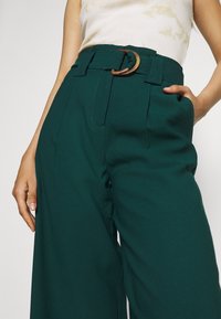 Teal vida byxor i mjukt tyg, med hög midja, framfåll och ett brunt runt spännebälte.