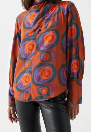 Vrouw draagt een loszittende blouse met lange mouwen met een oranje, paarse en groene cirkelvormige print, gecombineerd met zwarte leren broek.