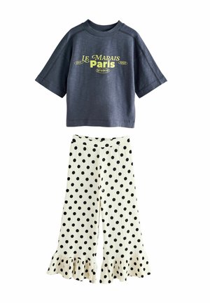 Granatowy krótki top z żółtym napisem "Le Marais Paris Studio", w połączeniu z białymi szerokimi spodniami w czarne kropki i falbankami na nogawkach.