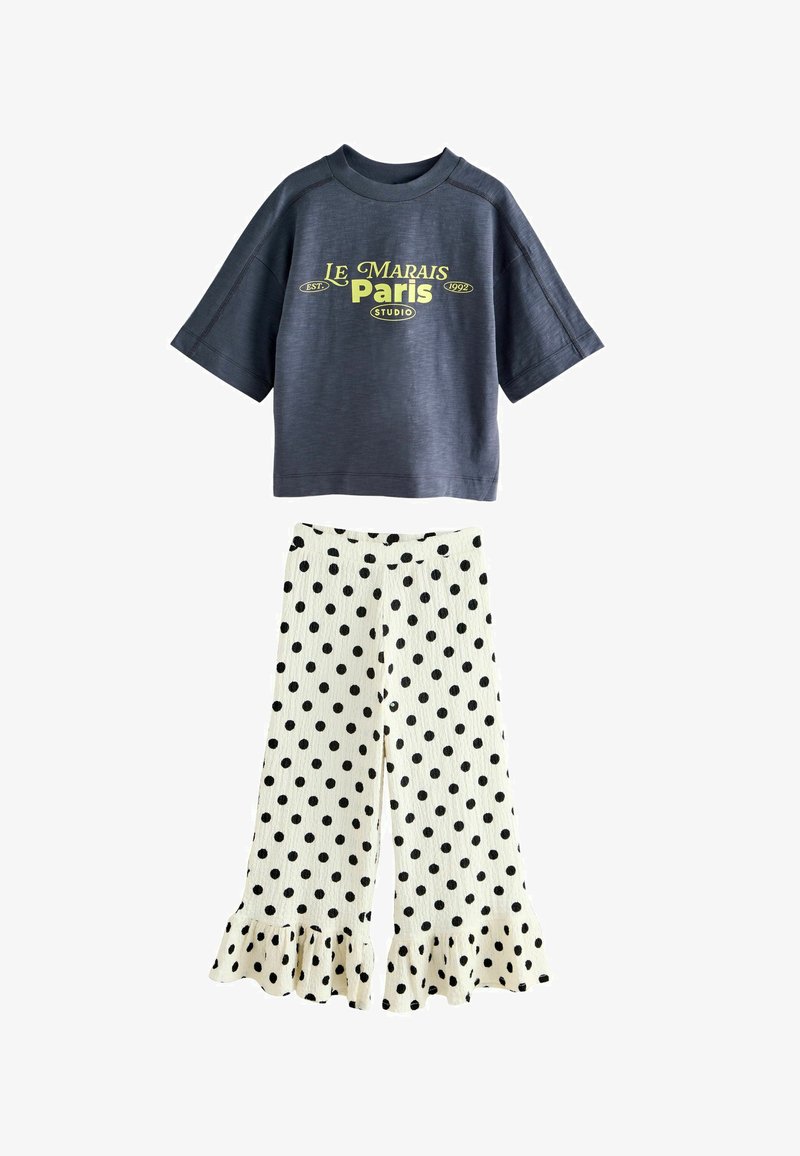 Temno modra majica s kratkimi rokavi z rumenim napisom "Le Marais Paris Studio", kombinirana z belimi širokimi hlačami z črnimi pikami in nabrano spodnjico.