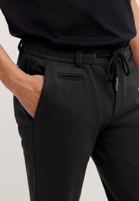 Hand in der Tasche einer schwarzen Hose mit Kordelzugbund, kombiniert mit einem schwarzen, hinein gesteckten Hemd.