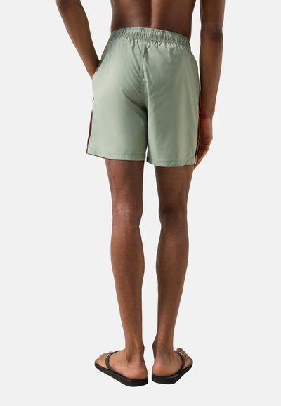 Personne debout pieds nus portant un short de bain vert clair avec des bandes latérales orange, photographiée de dos sur un fond uni.