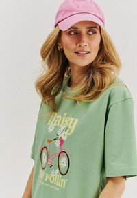 Zelené tričko s krátkymi rukávmi, so sladkým grafickým motívom ružového bicykla a kvetinovými vzormi, nosené s svetloružovou čiapkou. Vyrobené z mäkkého bavlneného materiálu.