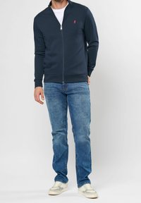 Marineblå zip-up sweatshirt med ståkrage, ribbade manschetter och nederkant; har en liten röd logotyp. Bärs med ljusblå jeans och krämfärgade sneakers.