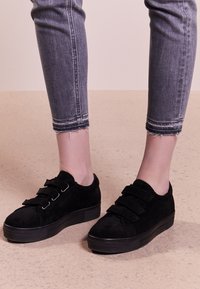 Zapatillas de ante negras con suelas gruesas; cuentan con tres correas de Velcro y un diseño de punta redondeada. Llevadas con jeans grises de dobladillo deshilachado.
