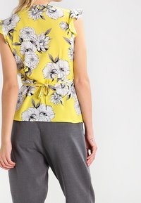 Donna che indossa un top senza maniche floreale giallo con spalline arricciate e pantaloni grigi, vista di spalle.