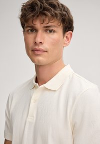 Weißes Poloshirt mit einem strukturierten Wellenmuster, ausgestattet mit einem klassischen Kragen und einer Drei-Knopf-Leiste. Glattes Material, kurze Ärmel.