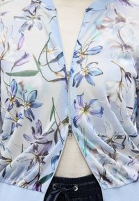 Ανοιχτό μπλε μπουφάν με φερμουάρ και μοβ και λευκά λουλουδάτα σχέδια, φορεμένο πάνω από μπεζ μπλουζάκι και μαύρο παντελόνι με κορδόνι.