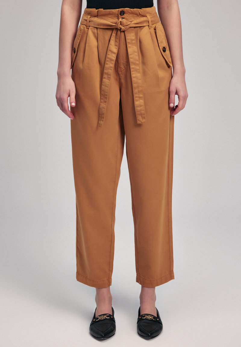 Tan hoge taillebroeken met een gestrikt riem, wijde pijpen, twee zijzakken en een gladde katoenen textuur. Draagt zwarte decoratieve flats.