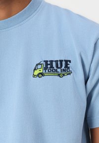 Light blue cotton T-shirt featuring a black and yellow embroidered logo of a truck with "HUF TOOL INC." text. Short-sleeved design.