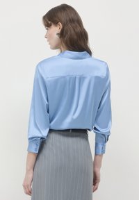 Camicia in satin azzurro chiaro con colletto, maniche lunghe e polsini abbottonati; presenta uno delicato yoke posteriore e una texture morbida. Abbinata a una gonna grigia a righe.