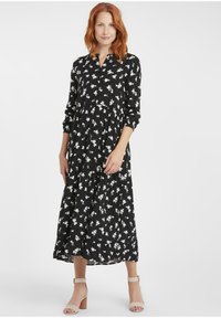 Schwarzes Kleid mit weißem Blumenmuster, langen Ärmeln, knopfbarem Kragen, gestuftem Rock, knöchellang, getragen mit beigen Absatzsandalen.