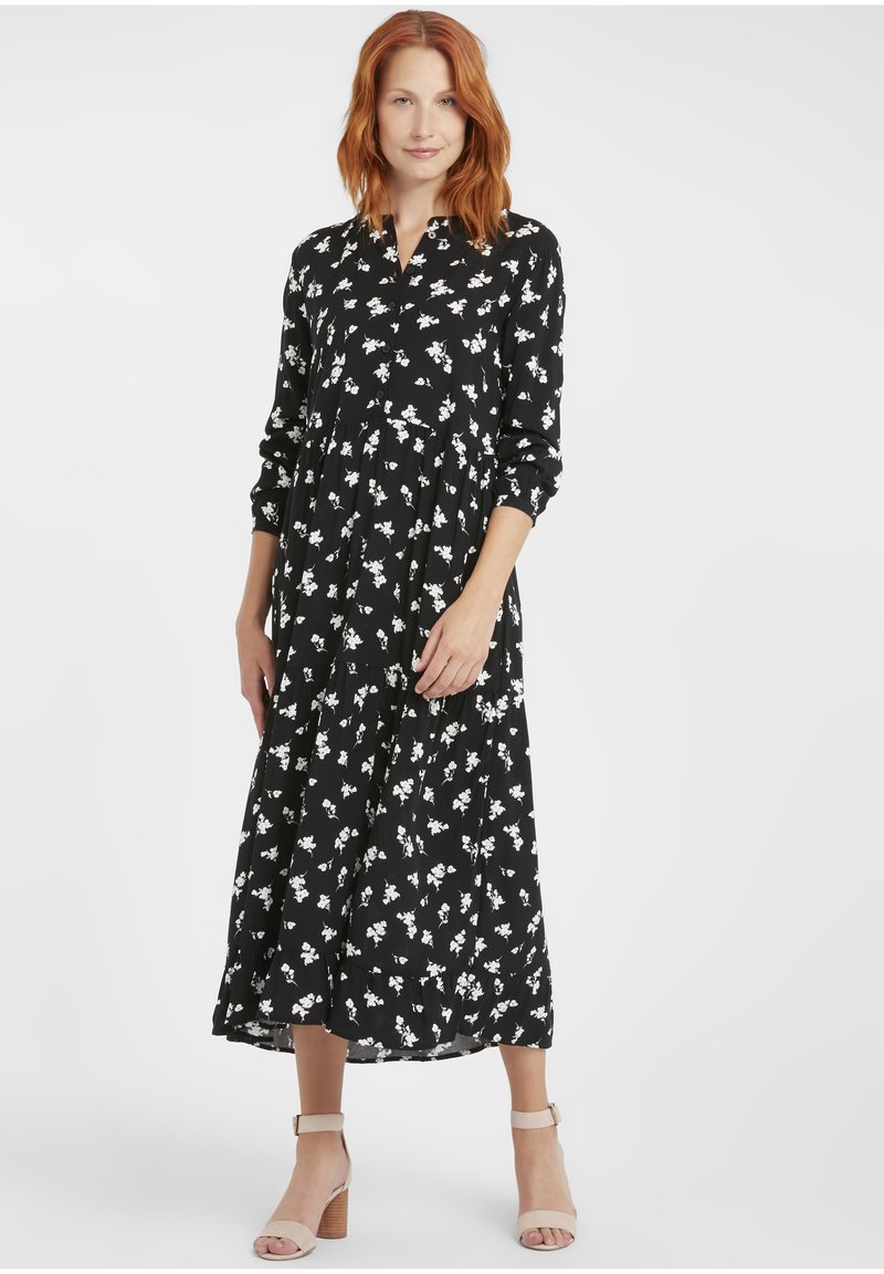 Schwarzes Kleid mit weißem Blumenmuster, langen Ärmeln, knopfbarem Kragen, gestuftem Rock, knöchellang, getragen mit beigen Absatzsandalen.