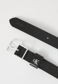 Juodas odinis diržas su sidabriniu stačiakampiu sagtimi ir diržo kilpa, kurioje iškeltu sidabriniu "cK" logotipu ant balto fono.