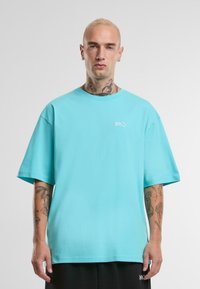 Turquoise oversized katoenen T-shirt met een ronde hals en korte mouwen, met een klein wit logo op de linkerborst. Soepele textuur.