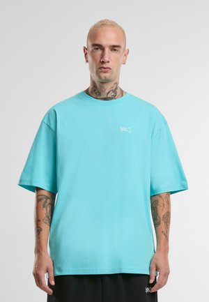 T-shirt en coton surdimensionné turquoise avec un col rond et des manches courtes, arborant un petit logo blanc sur la poitrine gauche. Texture lisse.