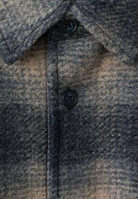 Vlnená kockovaná košeľa v odtieňoch šedej a béžovej, s golierom a tmavými gombíkmi, vyrobená z mäkkej, textúrnej látky a so štruktúrovaným dizajnom.