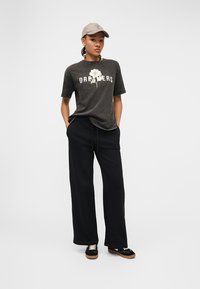 Νεαρή γυναίκα που φοράει γκρι μπλουζάκι με τη λέξη "Dreamers" και ένα λευκό λουλούδι, μαύρο παντελόνι με φαρδύ πόδι, μαύρα αθλητικά παπούτσια και μπεζ καπέλο, στέκεται με τα χέρια στις τσέπες.