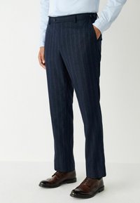 Tengerészen csíkos nadrág, struktúrált anyagból, testre szabott szabással, két elülső zsebbels és barna bőr cipővel viselve.