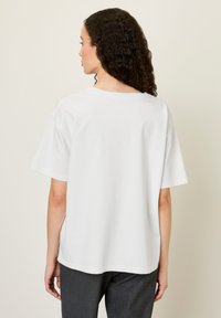T-shirt en coton blanc à manches courtes, avec un col rond et une coupe décontractée, associé à un pantalon gris foncé. La vue arrière met en évidence le design simple.