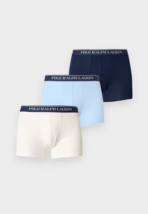 Muskulózny mužský model stojaci bosý v tmavomodrých Polo Ralph Lauren boxerkách na jednoliatom svetlom pozadí.