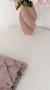 Rózsaszín bordázott kerámia váza művirágokkal, egy fehér asztalon elhelyezve, egy összehajtott virágmintás anyagról szóló műanyag táska mellett.