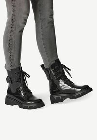 Schwarze Lackleder-Kampfstiefel mit einer massiven Sohle, Schnürung vorne, seitlichem Reißverschluss und dekorativem Schnallen-Detail, kombiniert mit grauen Skinny-Jeans.
