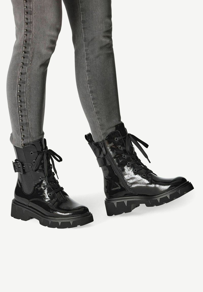 Schwarze Lackleder-Kampfstiefel mit einer massiven Sohle, Schnürung vorne, seitlichem Reißverschluss und dekorativem Schnallen-Detail, kombiniert mit grauen Skinny-Jeans.