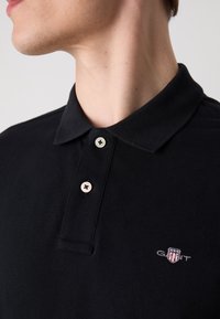 Black polo shirt with a classic collar, featuring two light-colored buttons and a small embroidered logo on the chest. Textured fabric.