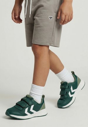 Dziecko noszące zielono-białe trampki na rzepy, białe skarpetki oraz beżowe szorty z logo pszczoły na nodze.