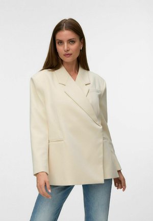 Femme aux longs cheveux bruns portant un blazer crème oversize et un jean bleu, debout devant un fond clair uni.