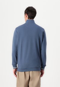 Μπλε φούτερ με κλασικό γιακά, ριχτά μανίκια με λευκές ρίγες και άνετη εφαρμογή. Λείος υφασμάτινος ιστός, μινιμαλιστικός σχεδιασμός.