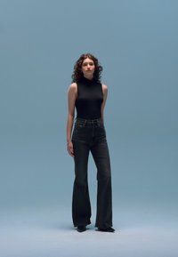 Top negro sin mangas de cuello alto, jeans acampanados de mezclilla oscura y zapatos negros, usados por una modelo contra un fondo azul claro.