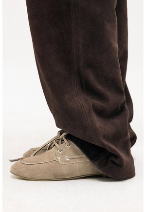 Hnedé semišové nohavice nosené nad svetlohnedými semišovými šnurovacími topánkami s dizajnom v štýle mokasínov na bielom pozadí.