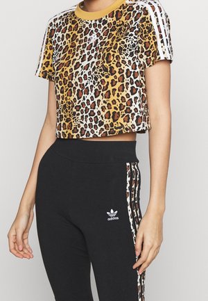 Femme portant un t-shirt court Adidas à imprimé léopard avec des rayures blanches et un legging Adidas noir avec des rayures léopard assorties.