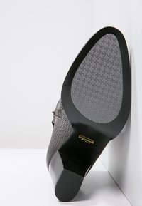 Scarpa grigia con texture e motivo a chevron sulla suola, dotata di tacco in gomma nera e dettagli metallici.