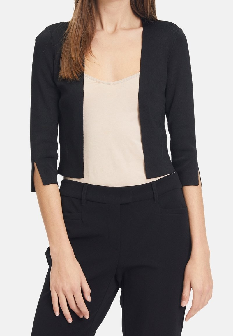 Black cropped cardigan, lightweight knit, open front design, three-quarter sleeves with side slits. Paired with a beige tank top and black pants.