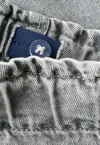 Hallhalli denimkangas, mis on halli värvi, sisaldab volangiga ääret ning sujuvat voldi. Näha on sinine tekstiilitagi ja läbipaistev plastiknööpide detail.