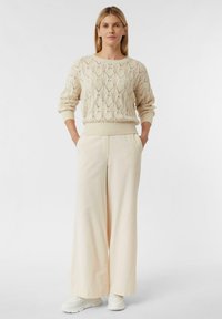 Strickpullover in hellem Beige mit rautenförmigen Mustern, kombiniert mit weit geschnittenen cremefarbenen Hosen, getragen mit weißen Sneakern. Glatte Textur insgesamt.