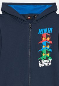 Nautička zip-up dukserica s plavom unutrašnjosti kapuljače. Sadrži grafiku triju ninja likova u crvenoj, plavoj i zelenoj boji, uz natpis "NINJA JAČI ZAJEDNO".