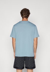 Lys blå bomull T-skjorte med korte ermer og rund hals, sett fra baksiden. Kombinert med mønstrete svarte shorts.
